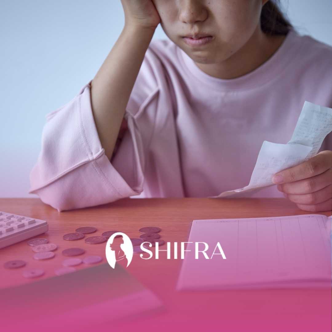 A person in a pink sweater sits at a table, looking worried while holding a receipt. Coins and a calculator are visible. The image conveys financial stress.