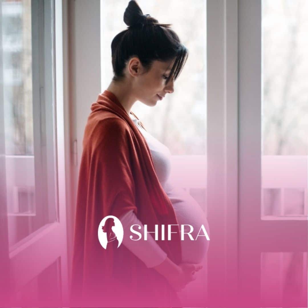 Pregnant woman looking down while standing by windows, overcoming stigma against premarital pregnancy.