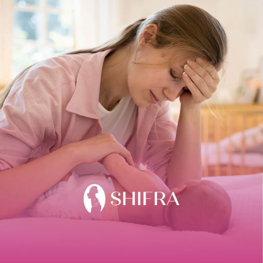 A tired mother, wearing a pink shirt, rests her forehead on her hand while holding a baby. The mood is weary yet tender.
